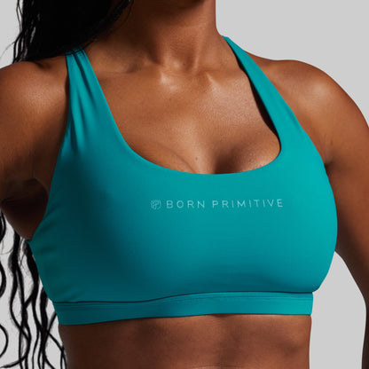 Teal sports bra with 'Born Primitive' logo worn by a person on a gray background