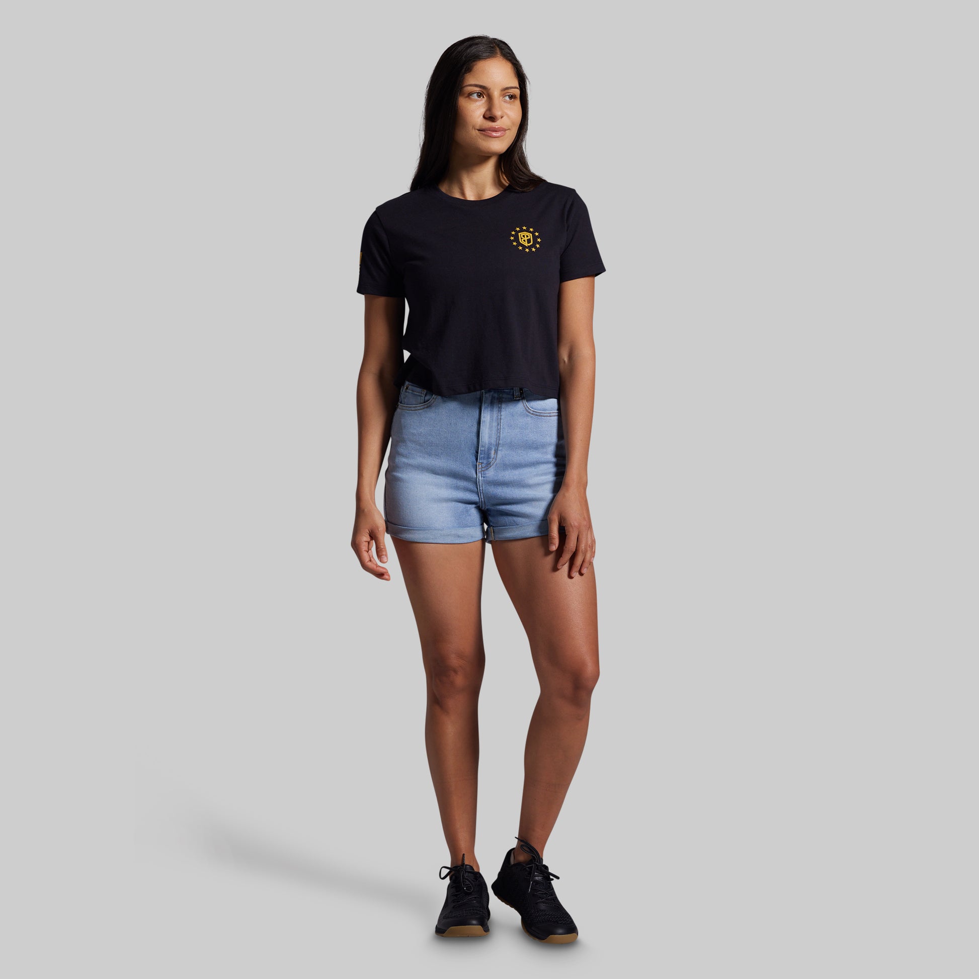 Woman wearing a black t-shirt with a yellow logo and denim shorts on a gray background