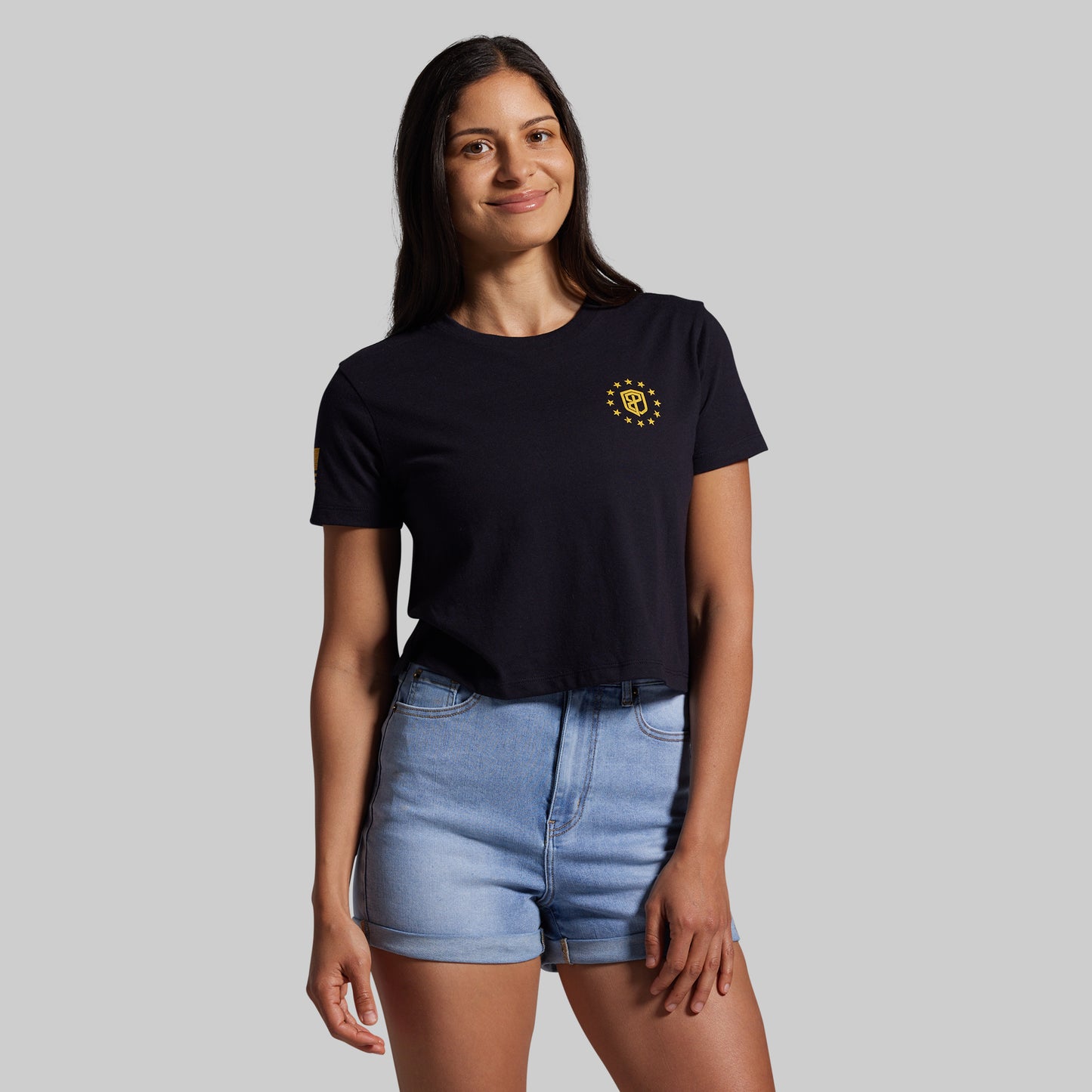 Woman wearing a black t-shirt with a yellow logo and light blue shorts on a gray background