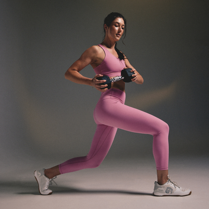A woman in a pink sports bra and matching leggings performs a lunge while holding a dumbbell, wearing white  sneakers against a dark gray studio background.