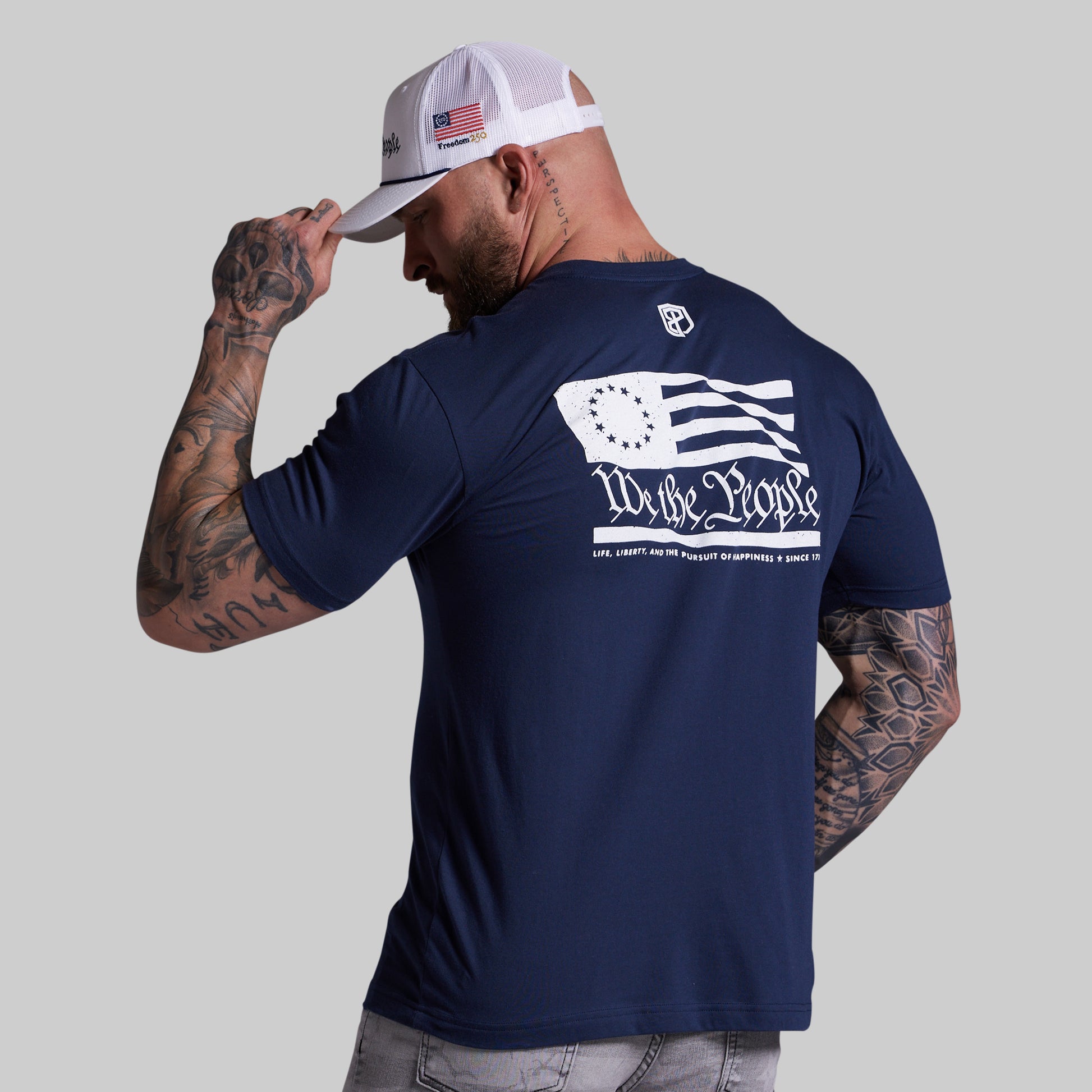 Man wearing a navy blue t-shirt with a flag design and text, tipping his cap against a gray background.