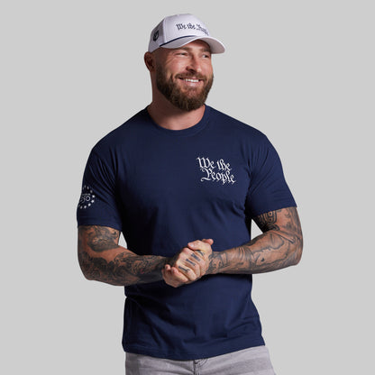 Man wearing a navy blue t-shirt with 'We the People' text and a white cap on a gray background
