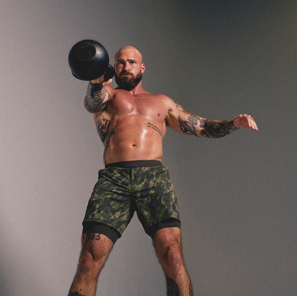A shirtless tattooed man holding a black kettlebell in a gym setting, wearing green and black camouflage athletic shorts against a plain gray background.