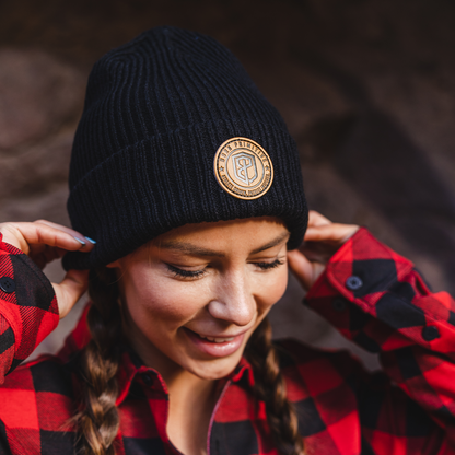A woman wearing a black ribbed knit beanie with a circular tan logo patch on the front, paired with a red and black buffalo plaid flannel shirt, set against a dark, blurred natural background.