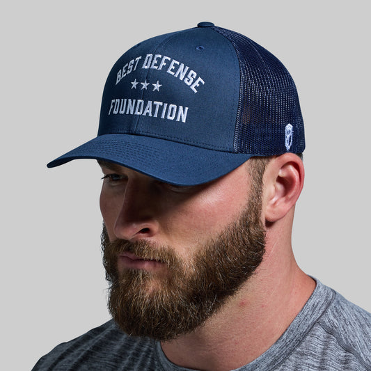 A person wearing a navy trucker hat with 'BEST DEFENSE FOUNDATION' text on the front.