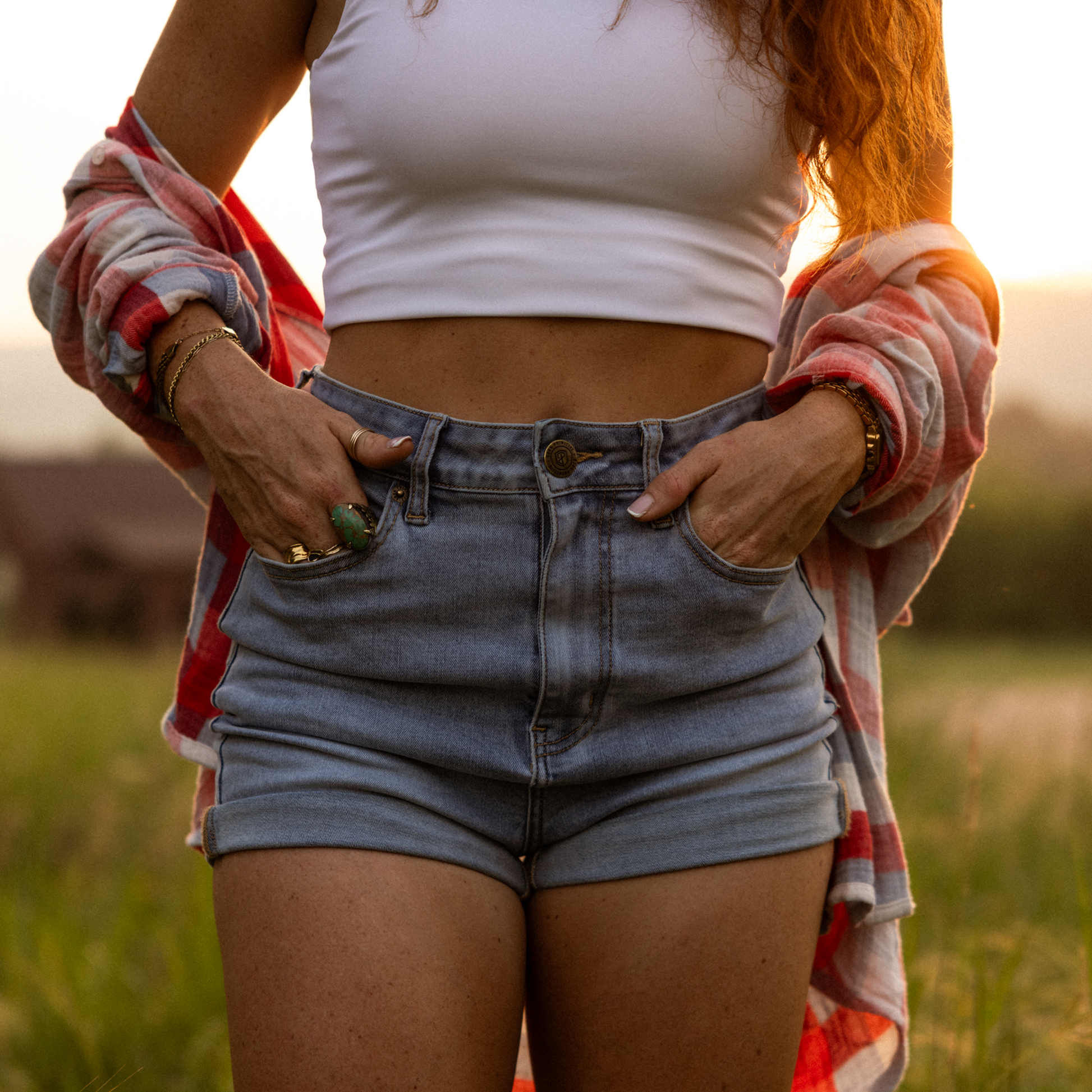 Person wearing a white crop top and blue shorts with a plaid shirt over shoulders in a field.