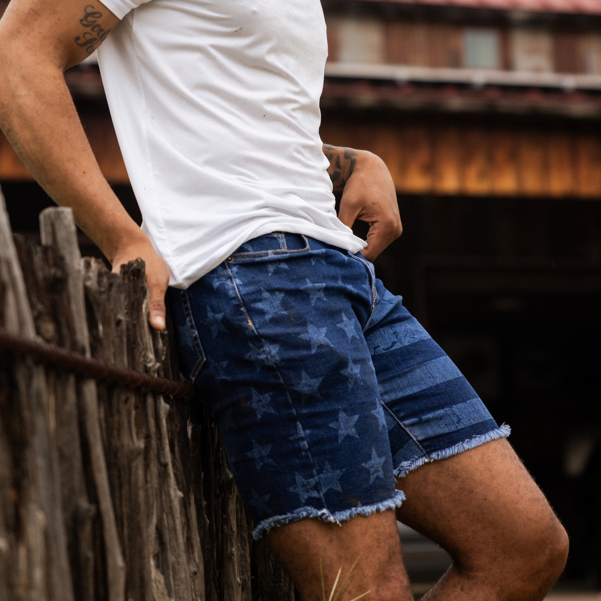 Person wearing a white t-shirt and blue denim shorts leaning against a wooden fence.