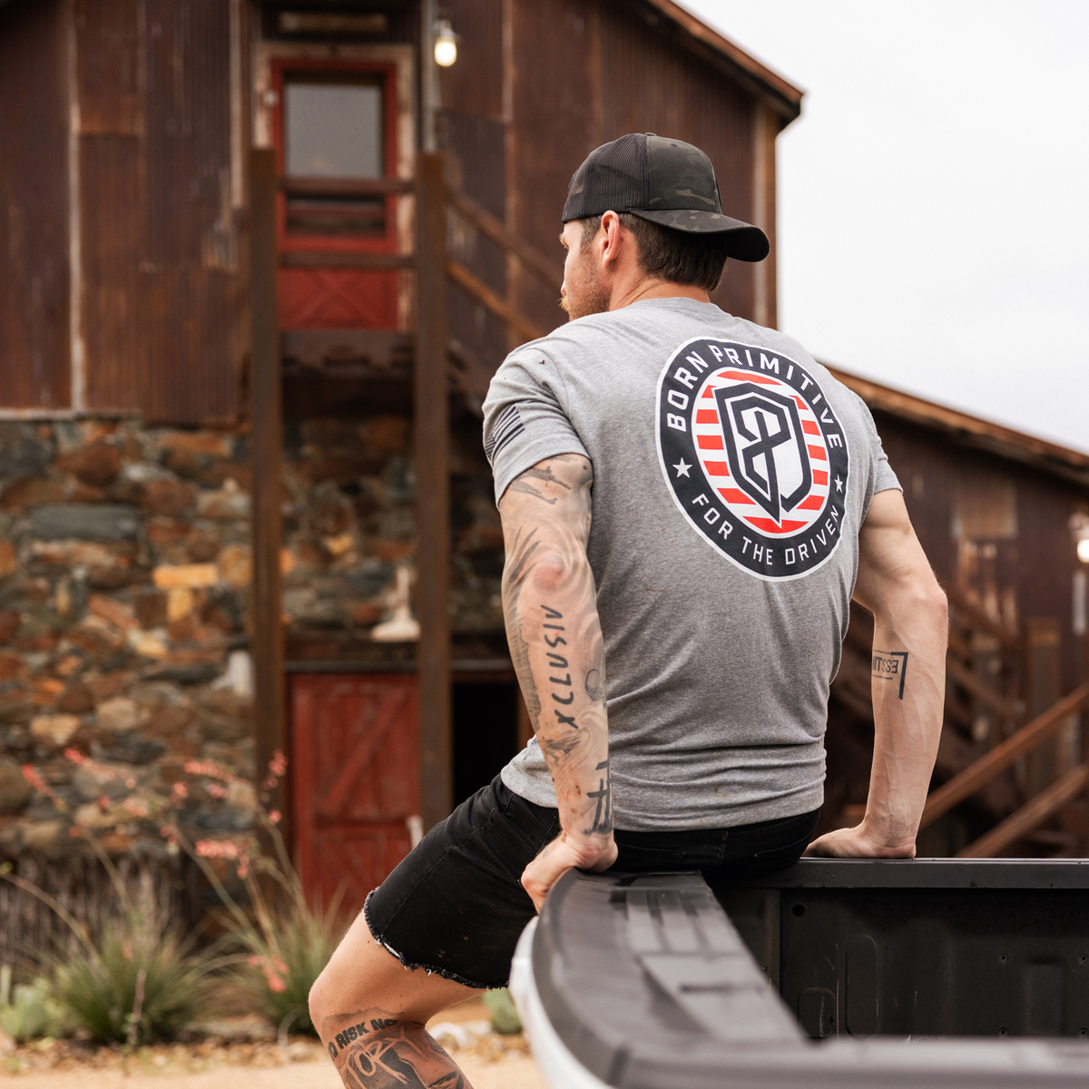 Man wearing a gray t-shirt with a logo, standing in front of a rustic wooden building.