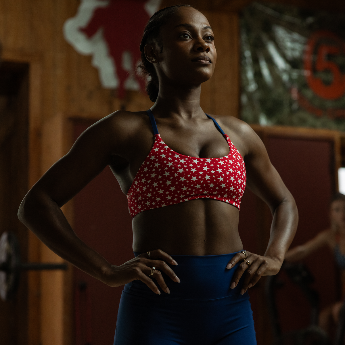 Woman in a red and white sports bra with stars and blue leggings standing in a gym.
