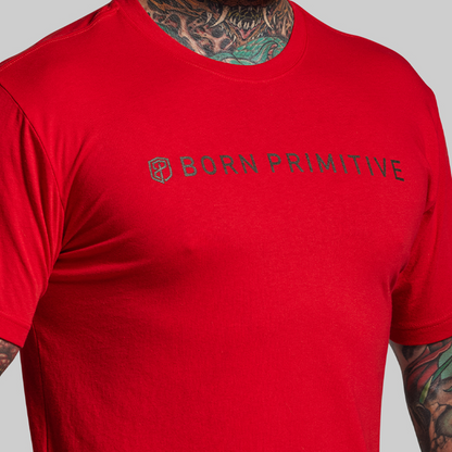 Red t-shirt with 'Born Primitive' logo worn by a person with tattoos on a gray background