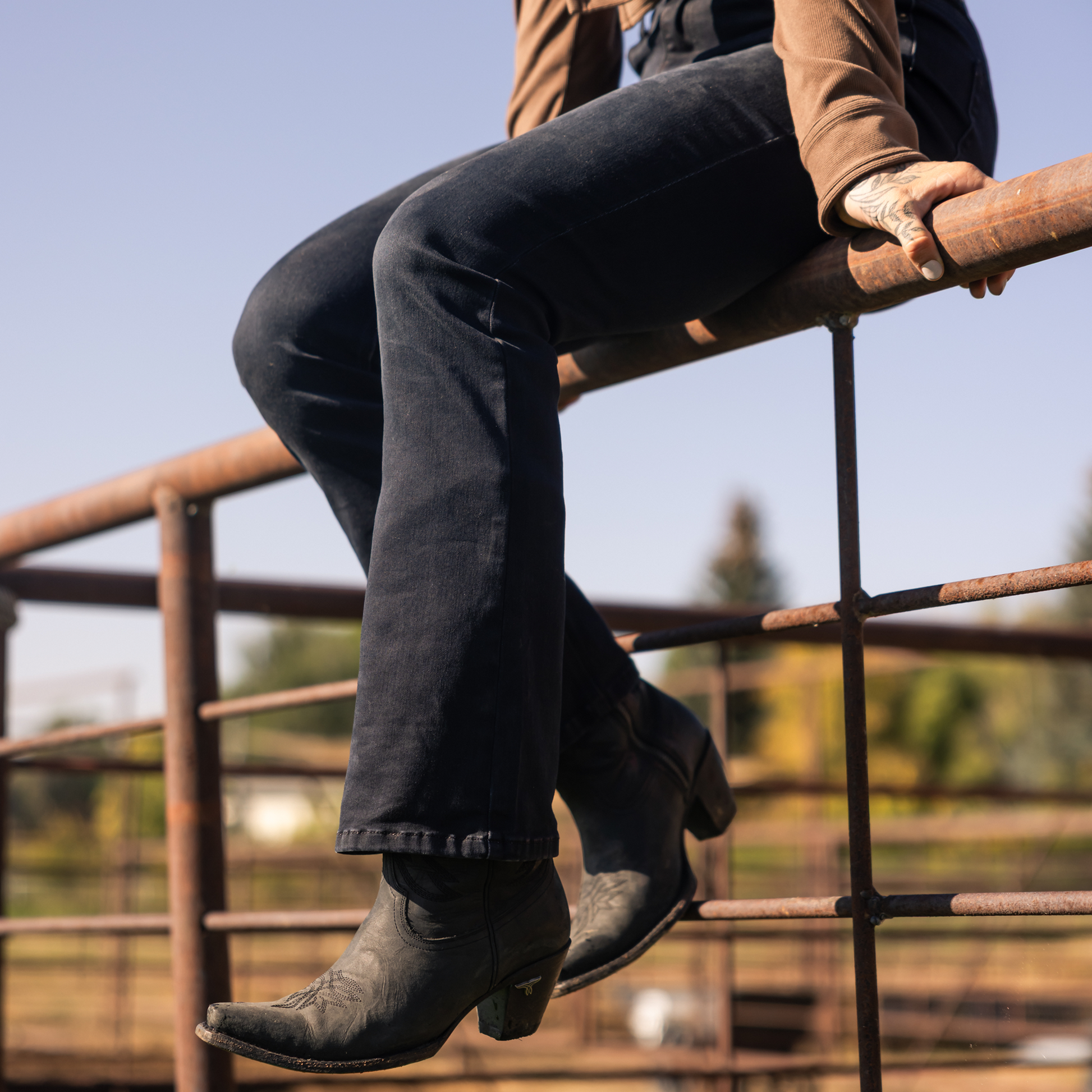Person wearing dark jeans and boots sitting on a metal fence with a blurred natural background