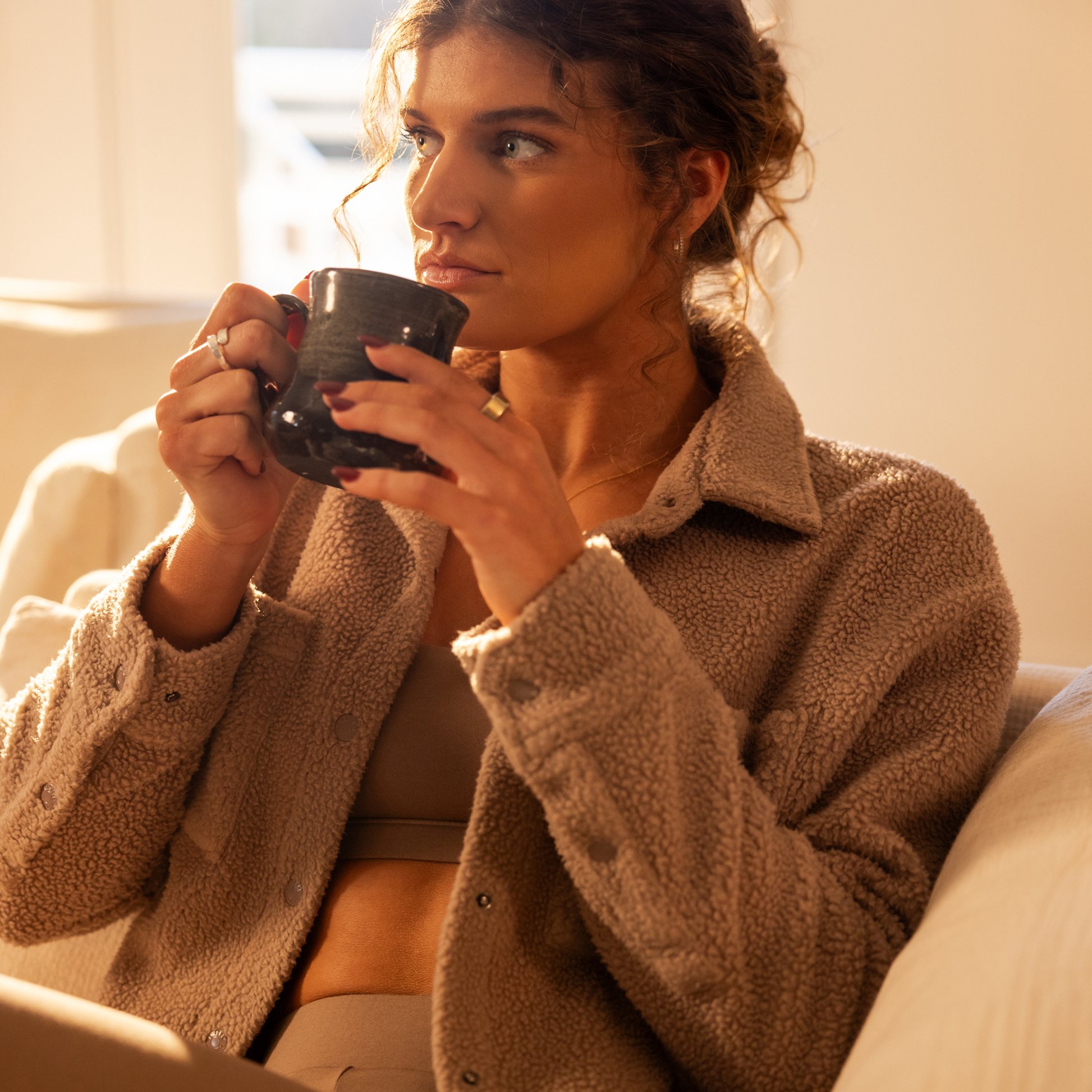 Woman in a cozy brown jacket holding a mug, sitting on a couch.