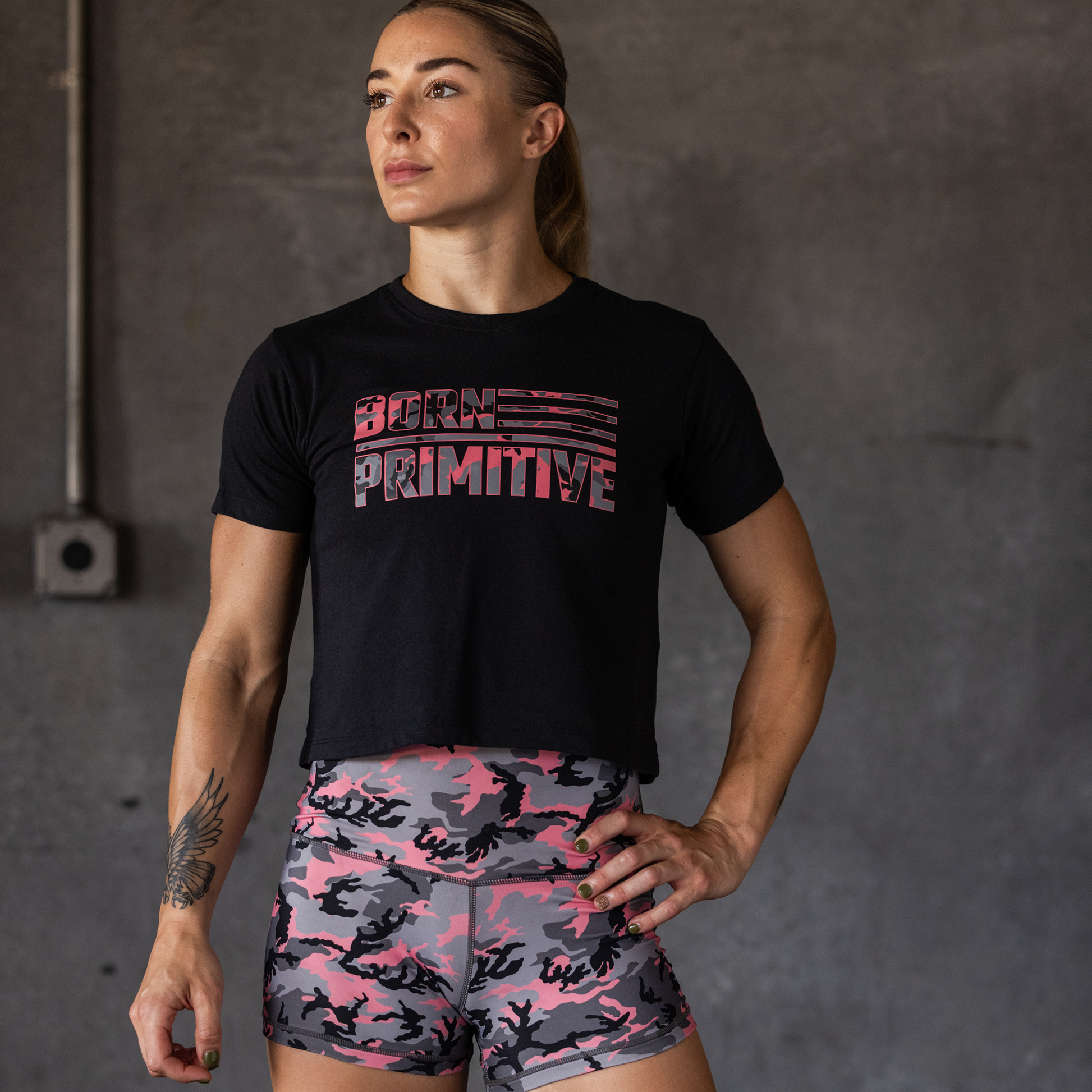 Woman wearing a black 'Born Primitive' t-shirt and pink camouflage shorts against a gray background