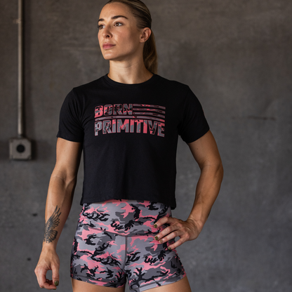 Woman wearing a black 'Born Primitive' t-shirt and pink camouflage shorts against a gray background