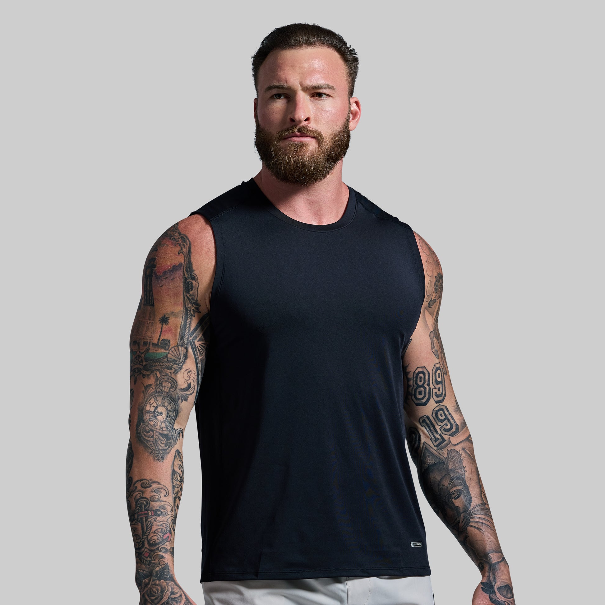 Man wearing a black sleeveless shirt with tattoos on arms against a plain background