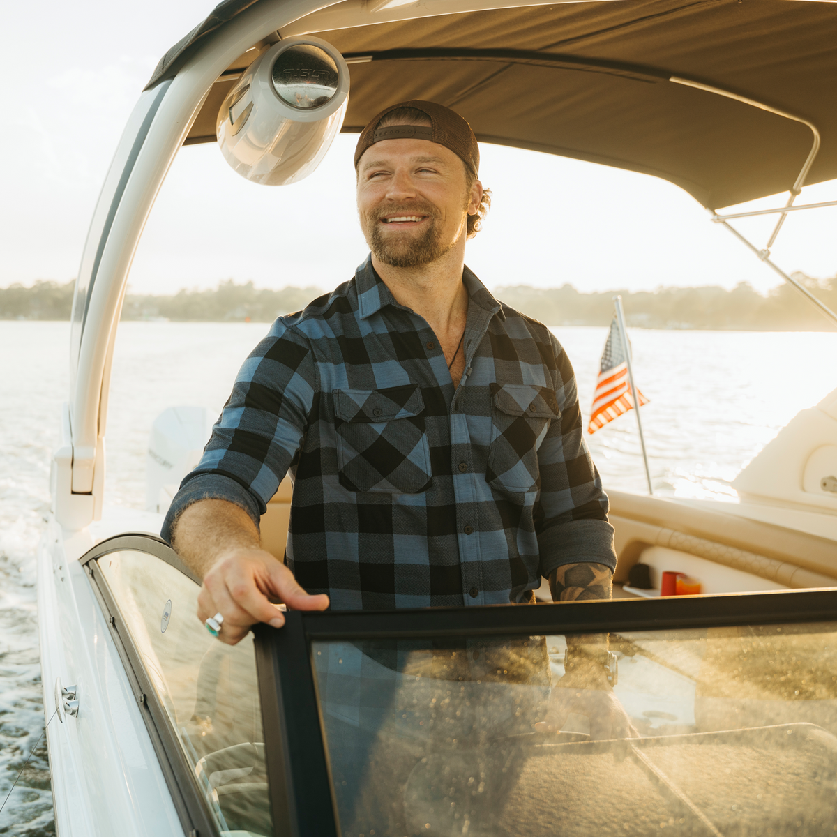 A smiling man wearing a blue and black plaid flannel shirt and a dark cap stands on a boat with water and trees in the background, holding the edge of the windshield.