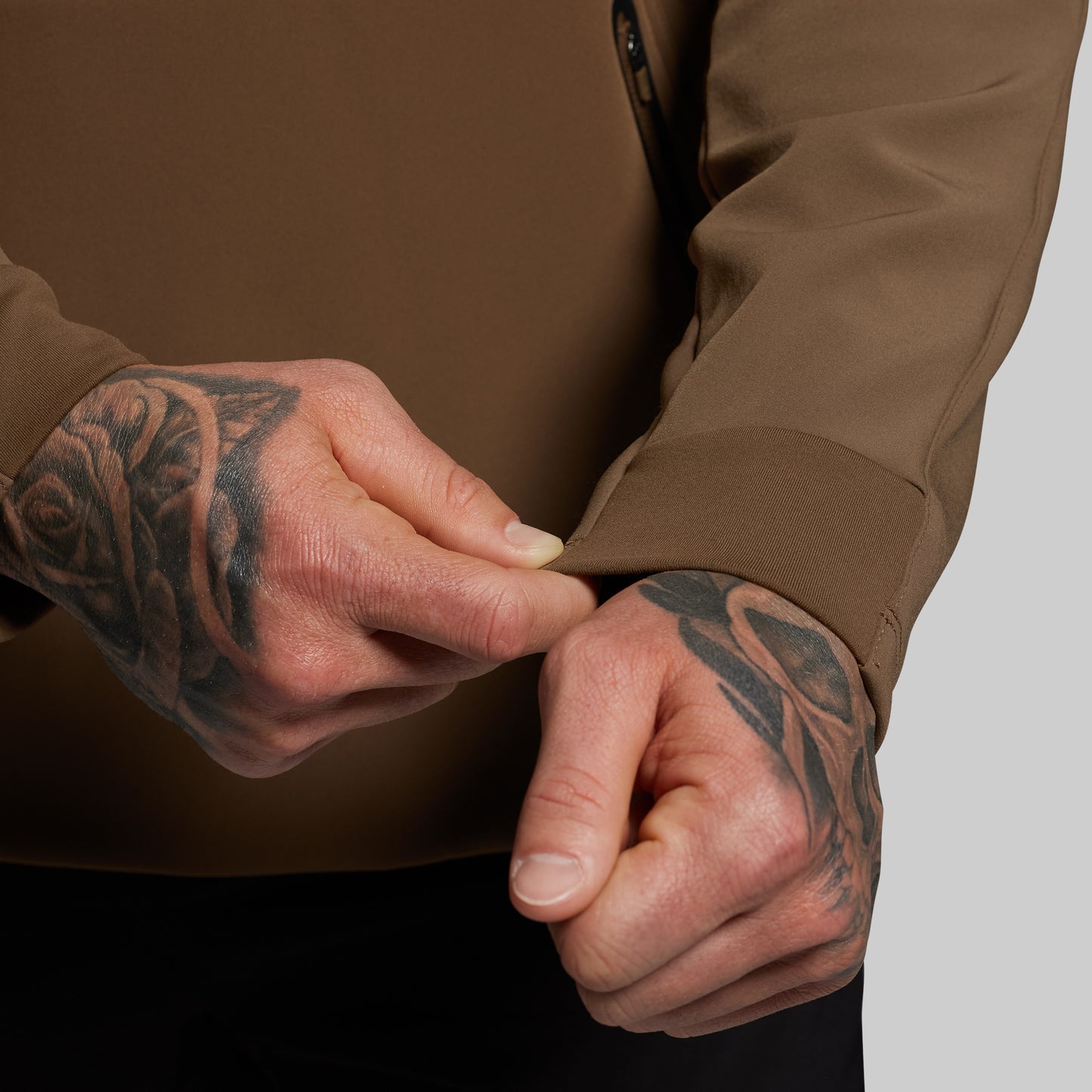Close-up of a person wearing a brown jacket with tattooed hands on a neutral background