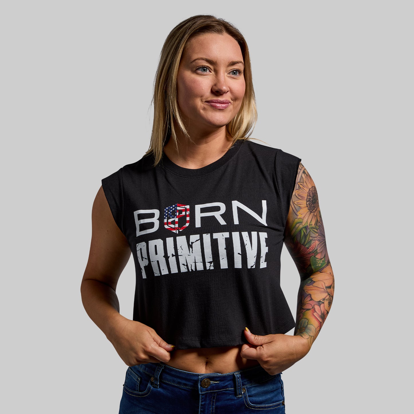 Woman wearing a black sleeveless shirt with 'Born Primitive' text, standing against a plain background