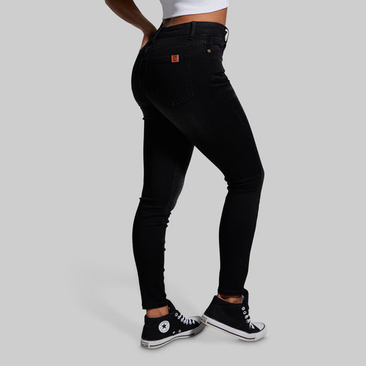 A model wearing black skinny jeans, shown from a side-back angle. The jeans feature a small orange brand patch on the back pocket.