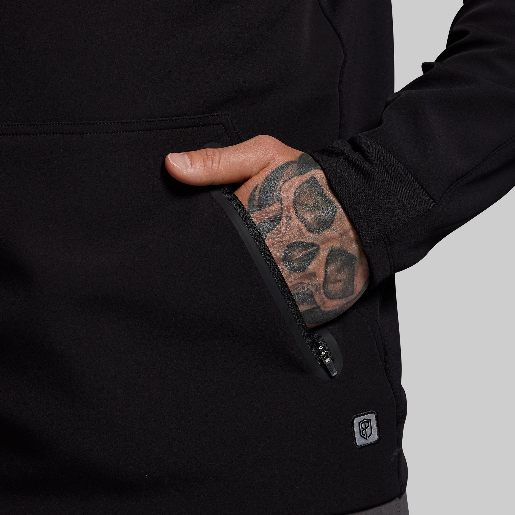 Person wearing a black jacket with a tattooed hand in the pocket on a gray background