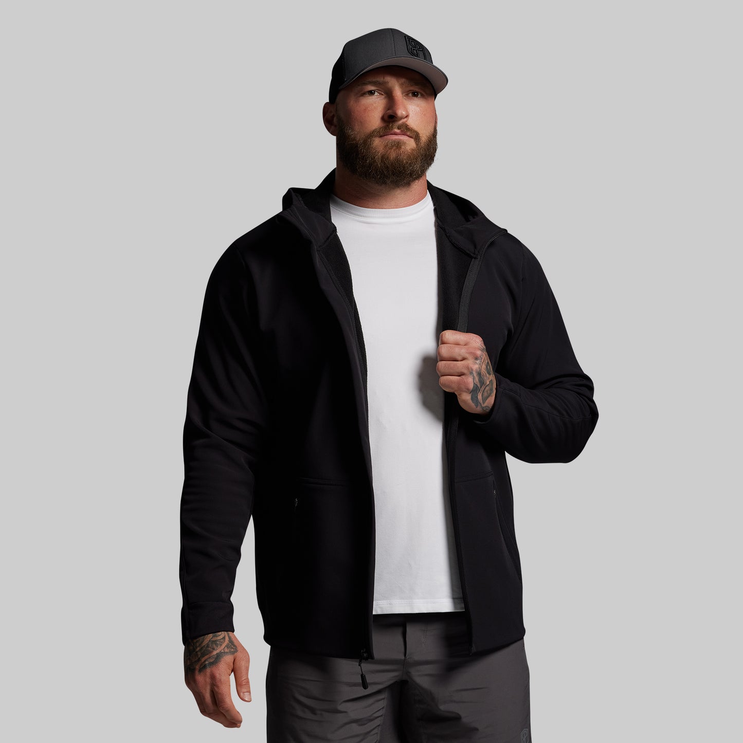 Man wearing a black full zip hoodie, white shirt, and cap on a gray background