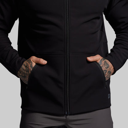 Person wearing a black jacket with tattoos on arms against a gray background