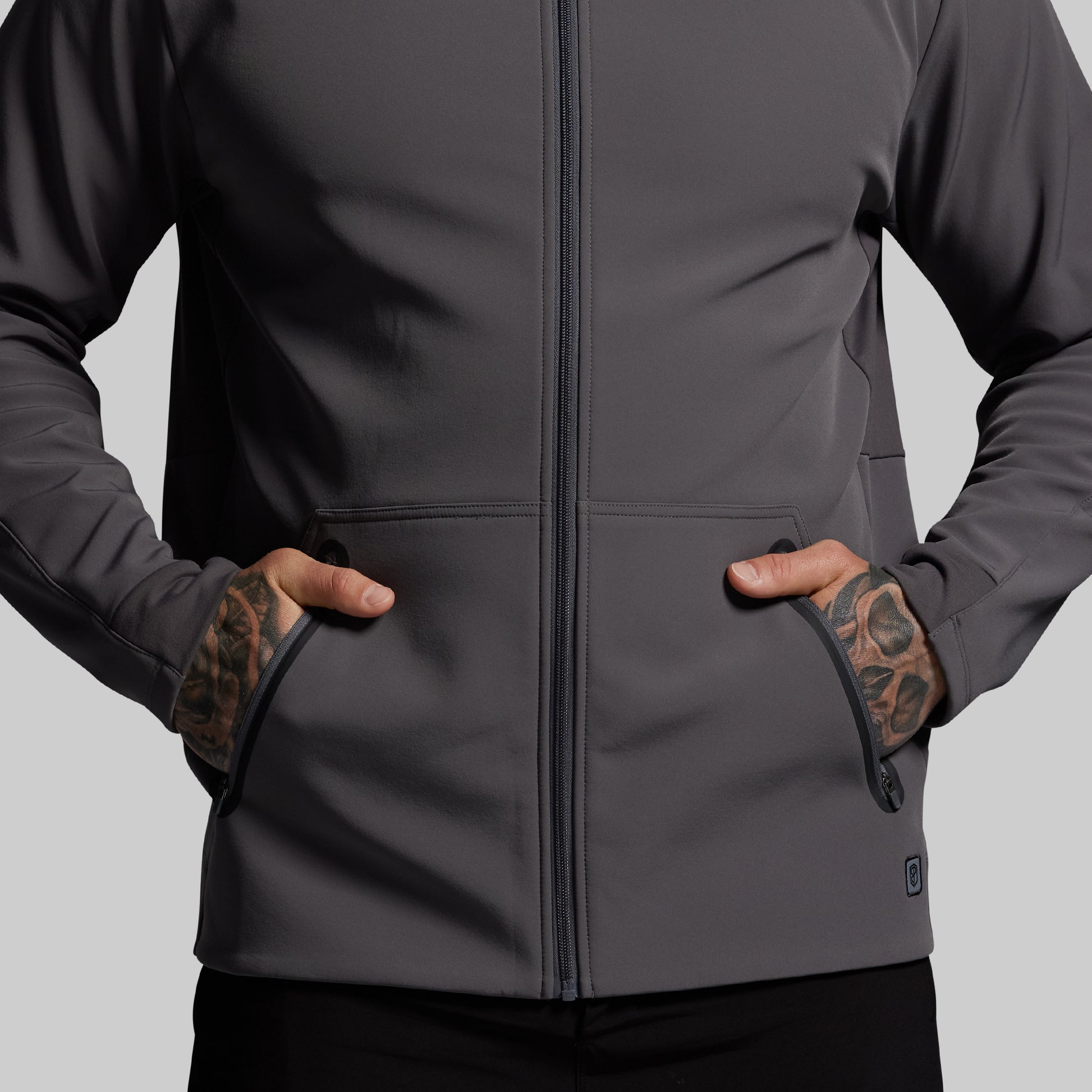 Person wearing a gray full zip hoodie with tattoos on arms against a light gray background