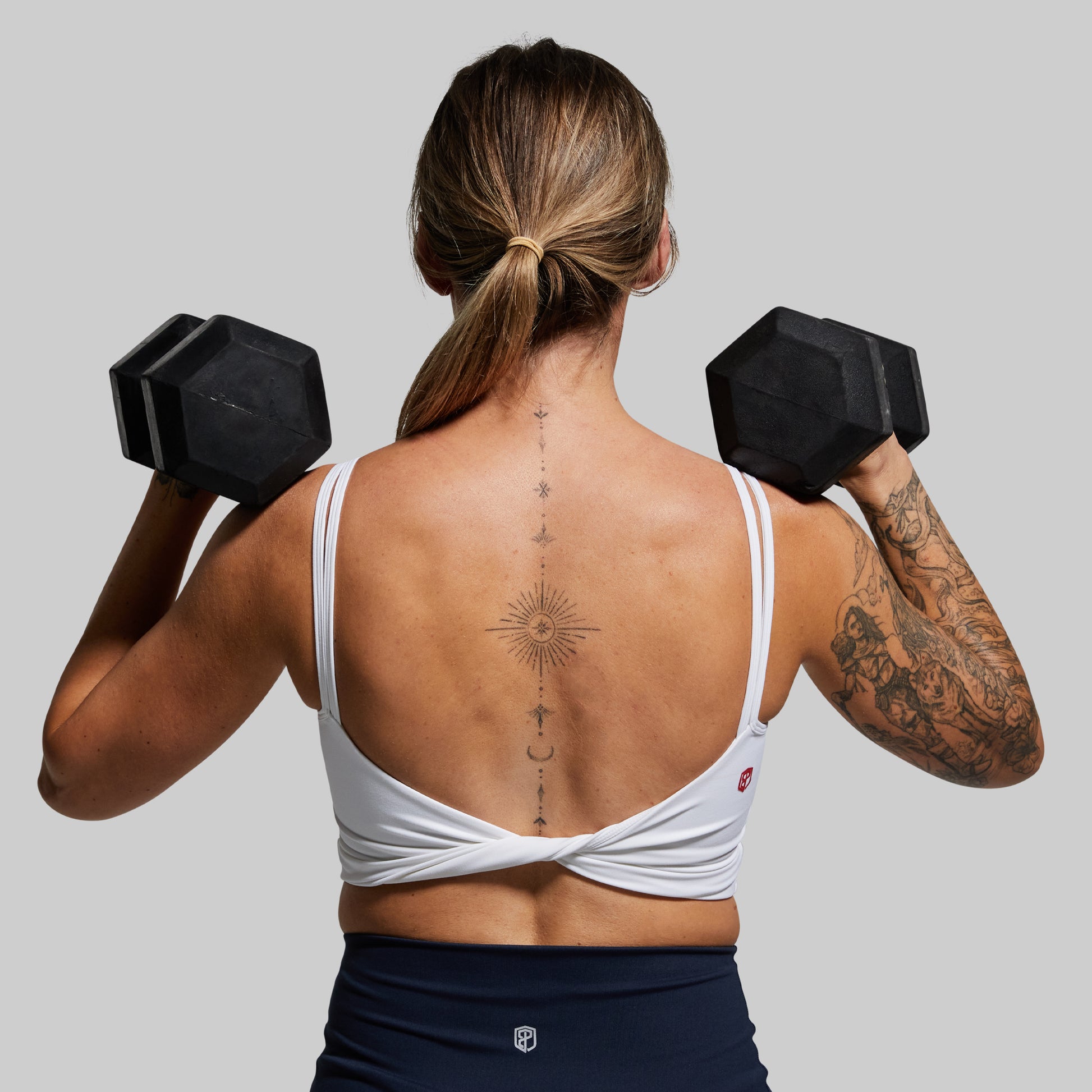 Woman lifting black dumbbells with tattoos on arms and back, wearing a white sports bra and dark pants.