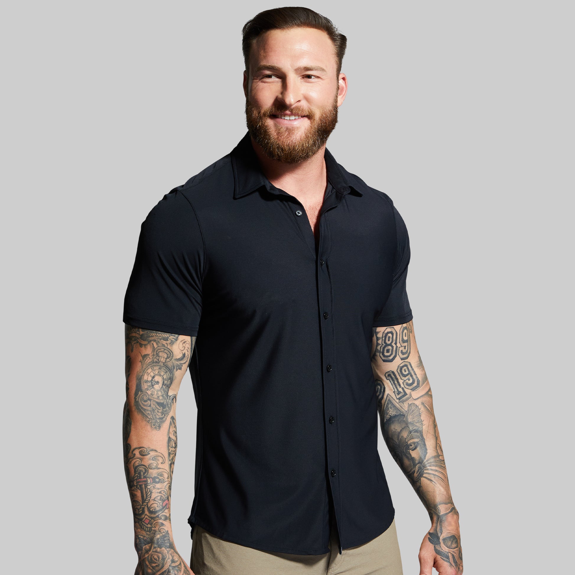 Man wearing a black short-sleeve shirt with tattoos on arms against a gray background