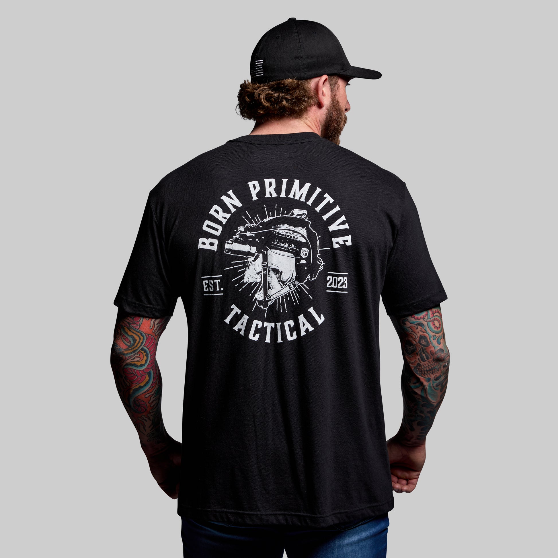 Man wearing a black 'Born Primitive Tactical' t-shirt with a graphic design on the back against a gray background