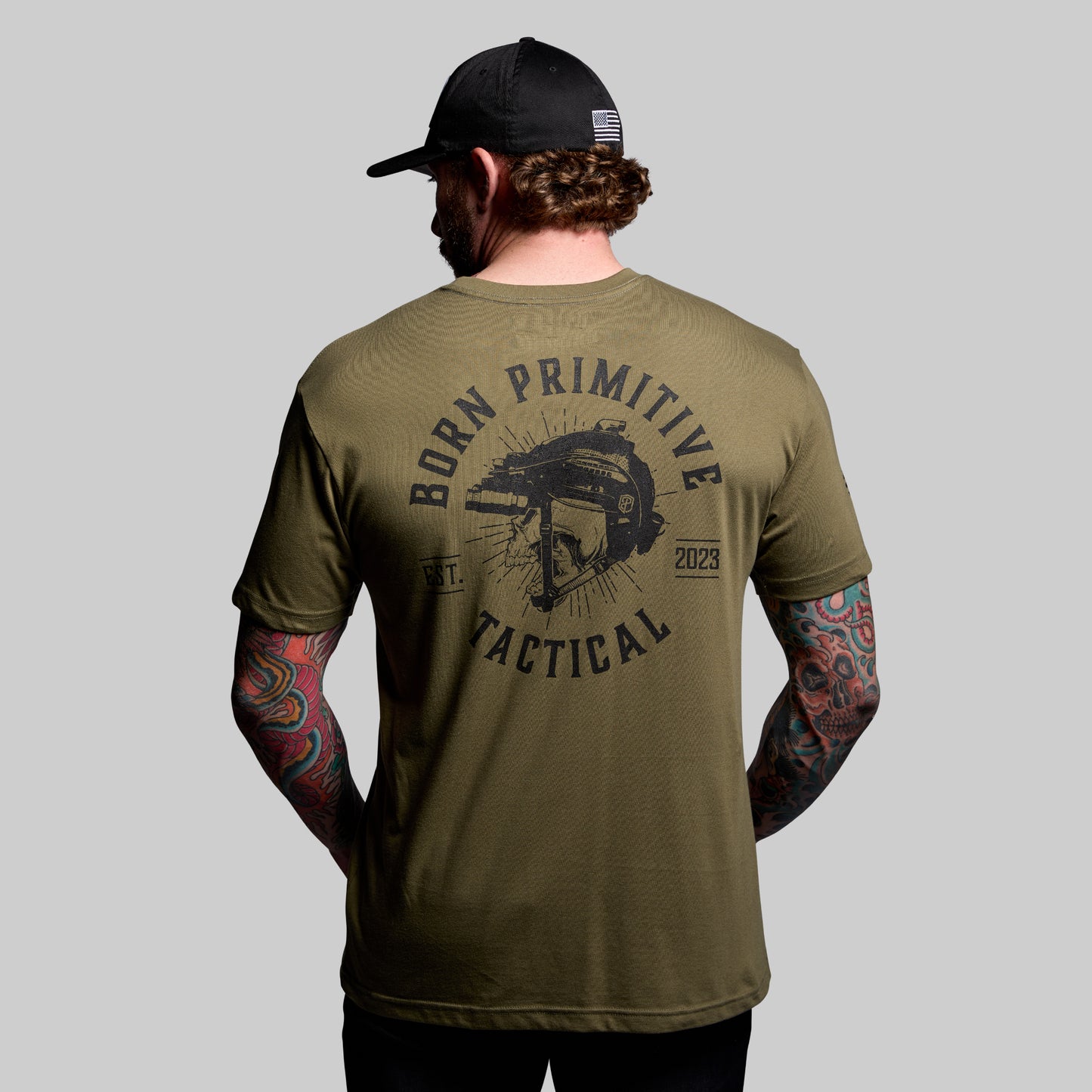 Man wearing a green 'Born Primitive Tactical' t-shirt with tattoos on arms, on a gray background