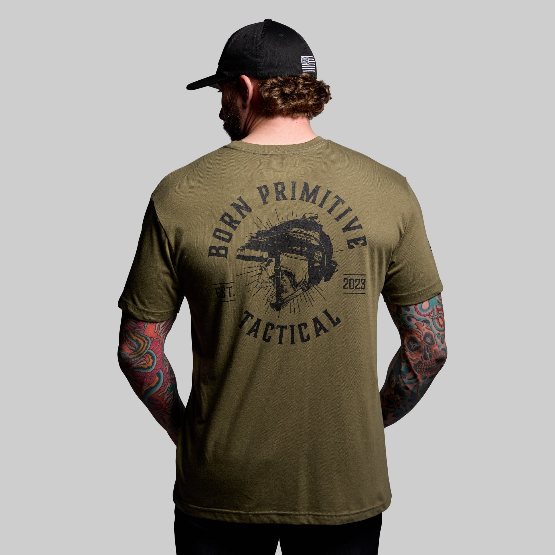 Man wearing a green 'Born Primitive Tactical' t-shirt with tattoos on arms, on a gray background