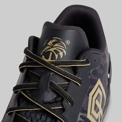 Close-up of a black athletic shoe with gold accents, featuring a gold eagle head logo on the tongue and a large gold geometric emblem on the side, set against a solid gray background.