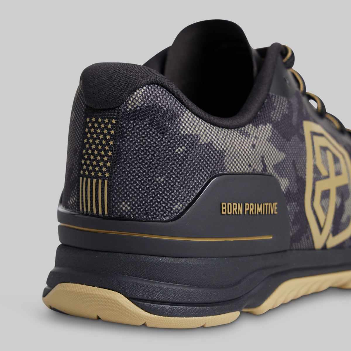 Close-up rear three-quarter view of a low-top athletic shoe in dark gray and black with gold accents, including a gold American flag motif on the heel, gold 'BORN PRIMITIVE' text, and a gold side logo, on a light gray background.