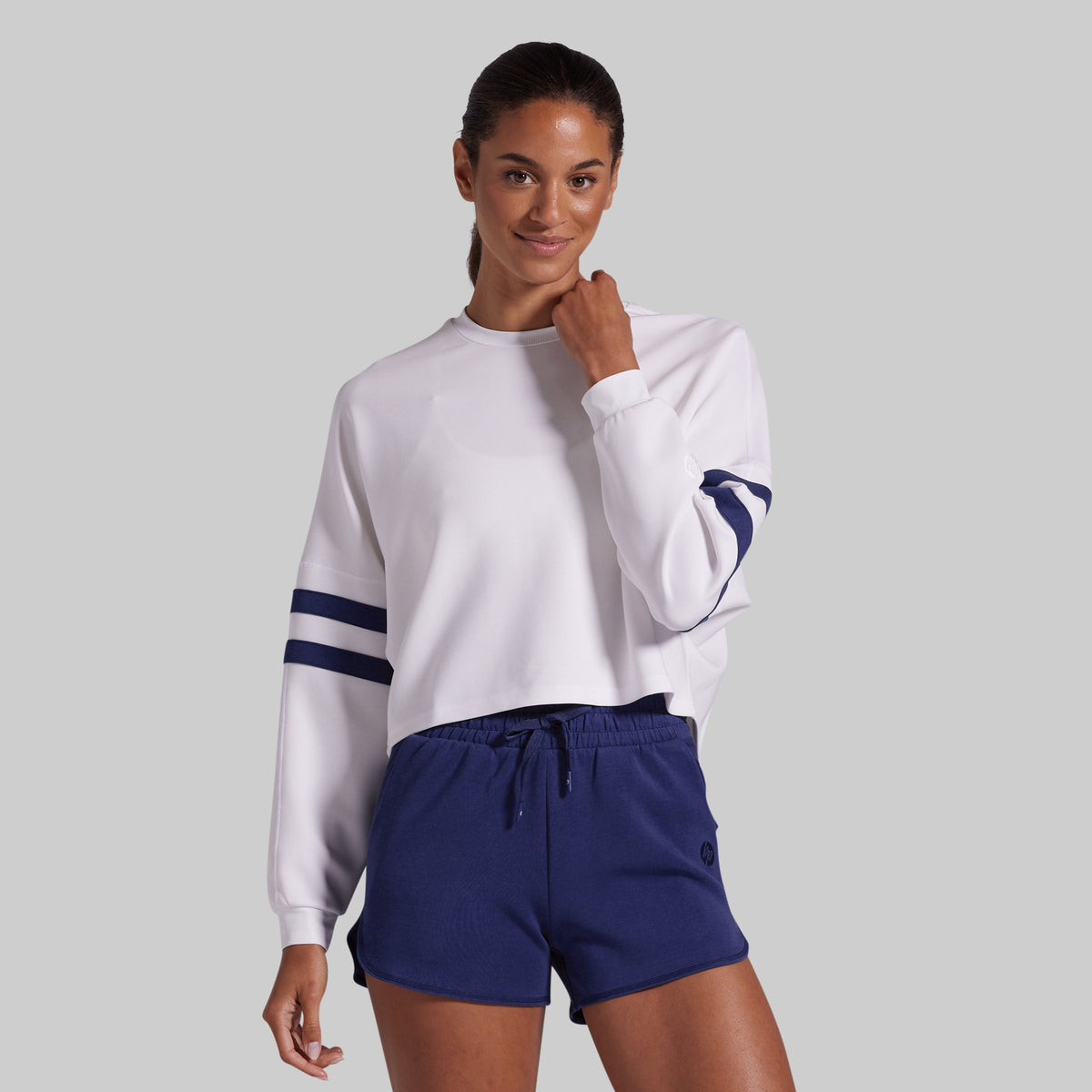 A woman modeling a white long-sleeve cropped sweatshirt with navy blue stripes on the sleeves, paired with navy blue lounge shorts, against a solid gray background.