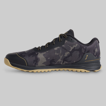 A low-top athletic shoe shown in a side profile on a solid gray background. The shoe features a dark gray and black camouflage pattern upper, black laces, a black toe cap, and a light tan outsole.
