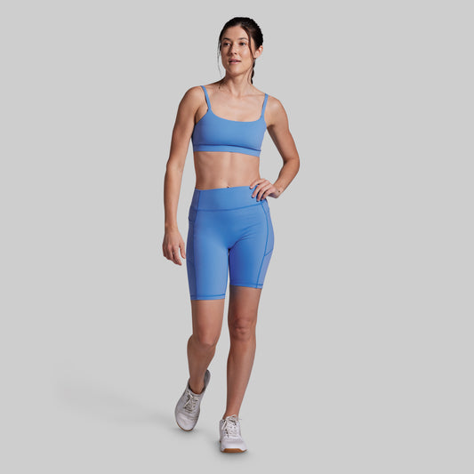 A woman modeling a matching athletic set consisting of a light blue sports bra and high-waisted biker shorts, paired with white sneakers, against a solid gray studio background.