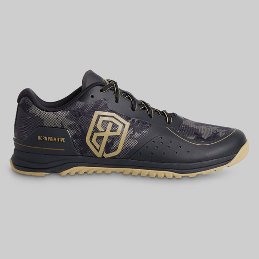 A single Born Primitive athletic shoe in black and dark gray camouflage with gold accents, including a large geometric logo on the side and 'BORN PRIMITIVE' text, shown against a solid gray background.