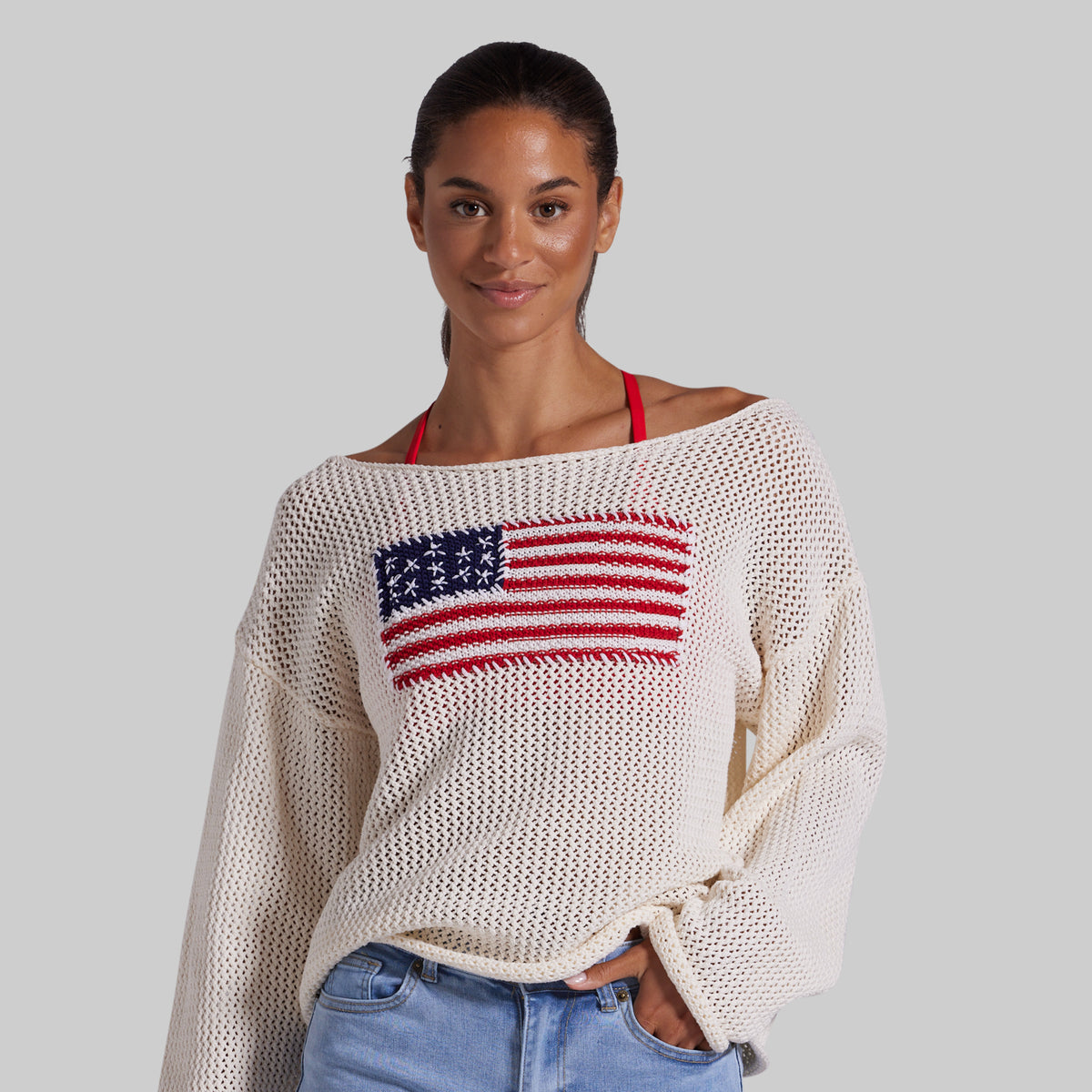 A woman wearing a cream-colored, off-the-shoulder knit sweater with a red, white, and blue American flag graphic on the chest, paired with light blue jean shorts, against a solid gray background.