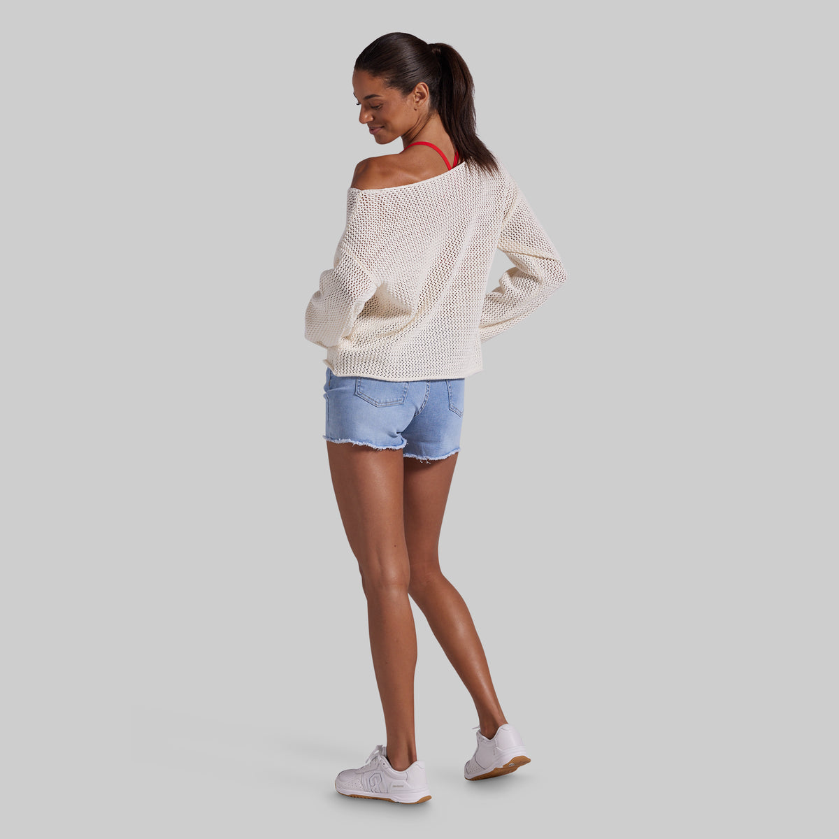 A woman modeling an off-the-shoulder cream knit sweater, light blue denim shorts, and white sneakers, shown from a side-back angle against a solid gray background.