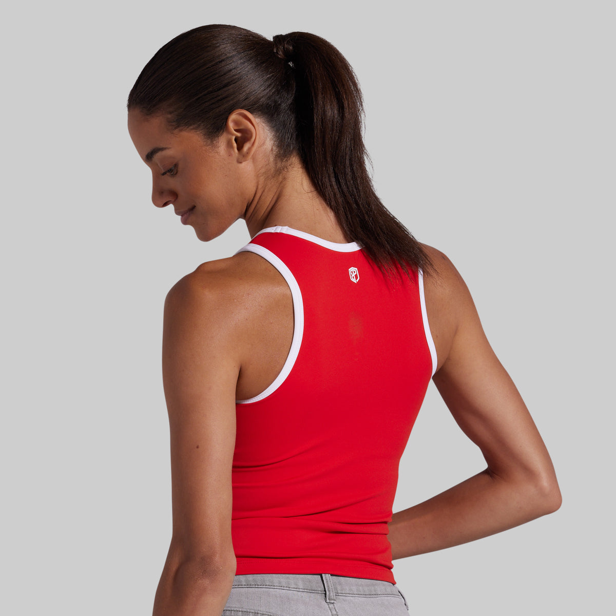 A woman wearing a bright red athletic tank top with white trim, shown from a side-back angle against a solid gray studio background.