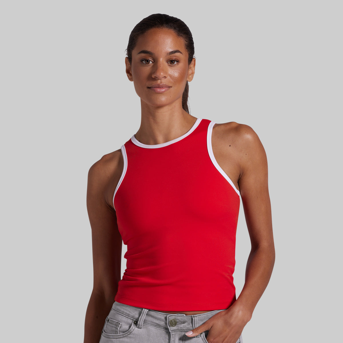 A woman modeling a bright red tank top with white trim along the neckline and armholes, paired with light gray jeans, against a solid light gray background.