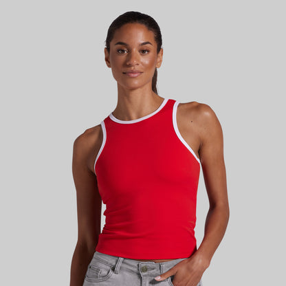 A woman modeling a bright red tank top with white trim along the neckline and armholes, paired with light gray jeans, against a solid light gray background.