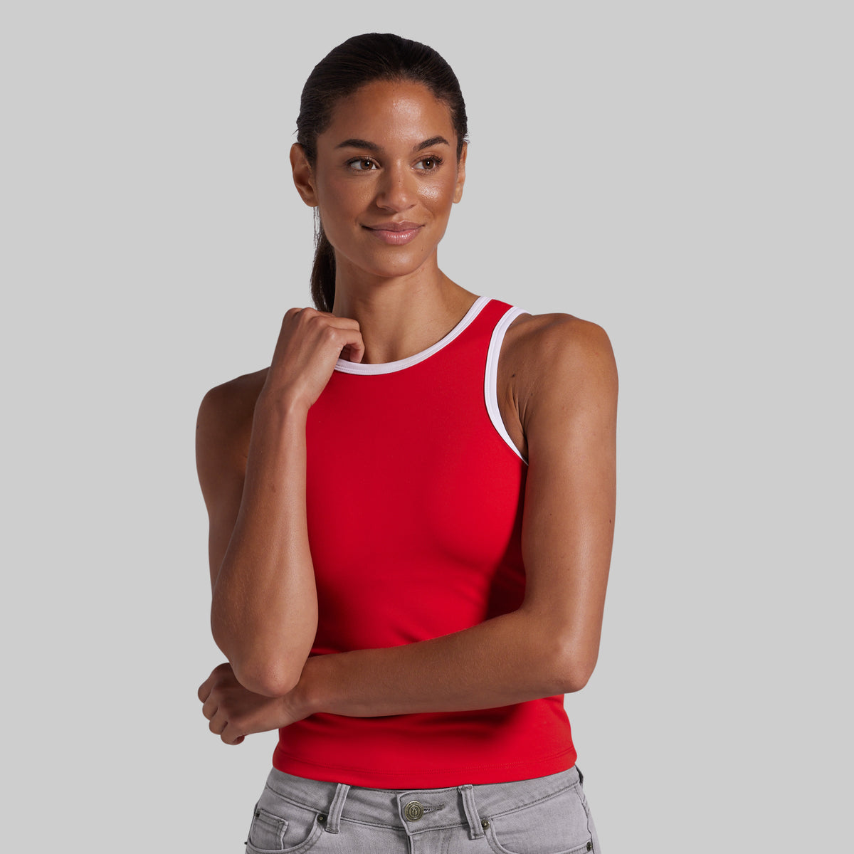 A woman modeling a bright red tank top with white contrast trim around the scoop neck and armholes, paired with light gray jeans, against a solid light gray background.