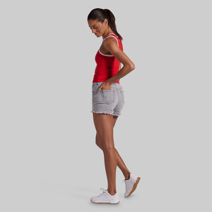 A woman in a red tank top, light gray denim shorts, and white sneakers is shown in profile against a solid light gray background.