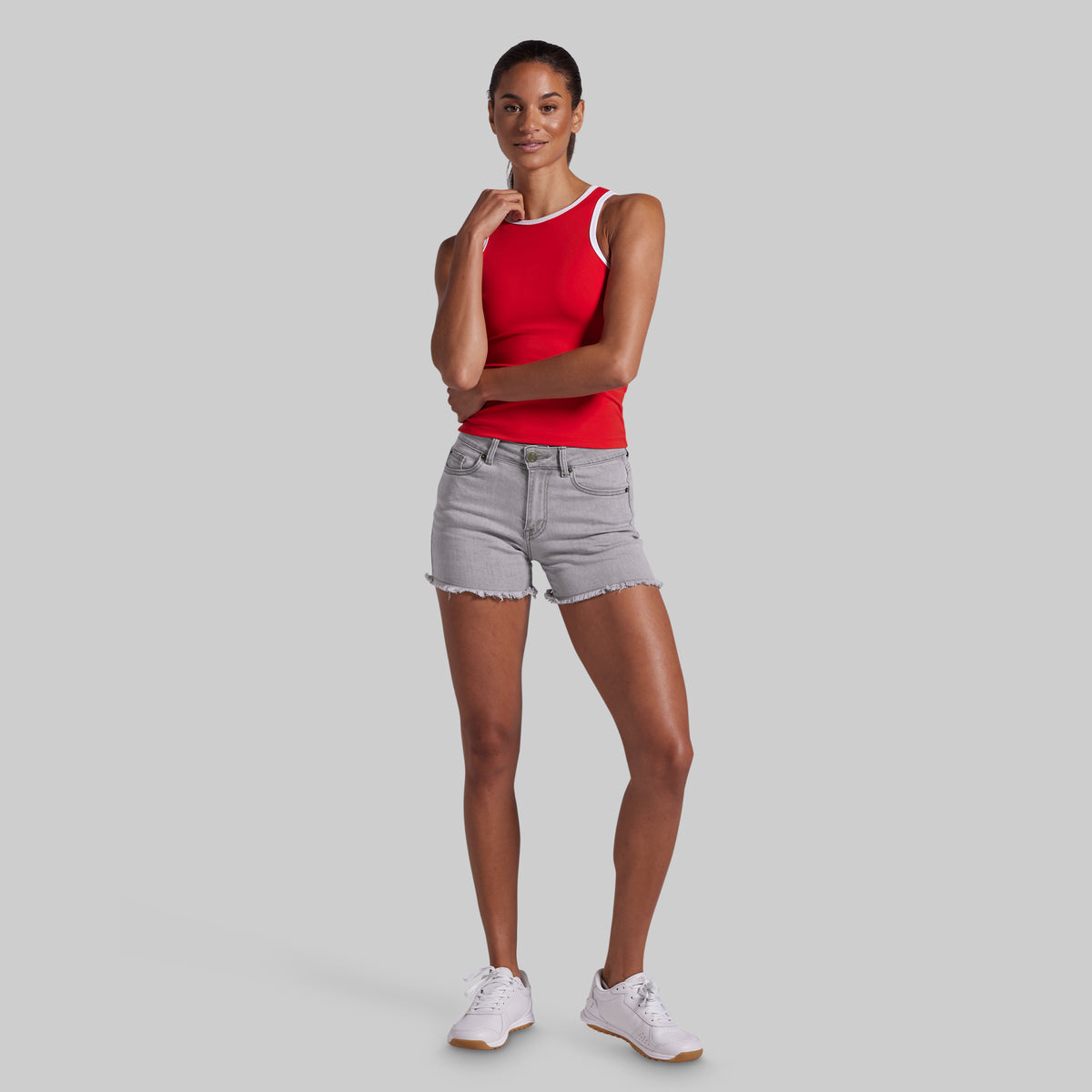 A woman modeling a bright red tank top with white trim and light gray denim shorts, paired with white sneakers, against a solid light gray background.