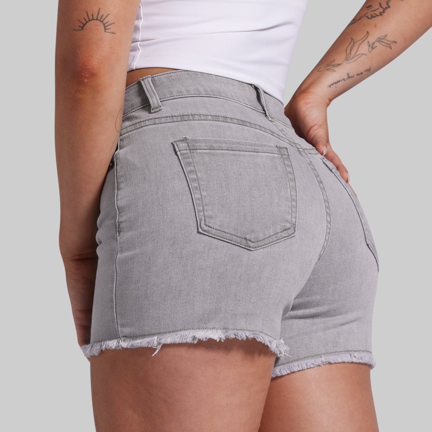 A close-up back view of a model wearing light gray denim shorts with a frayed hem and a back pocket, paired with a white top against a solid gray background.
