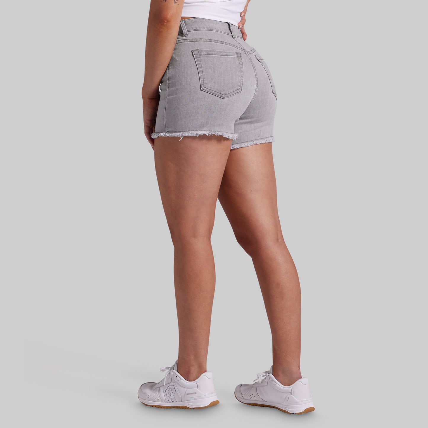 A person shown from the back wearing light gray frayed-hem denim shorts, a white top, and white sneakers against a plain light gray studio background.