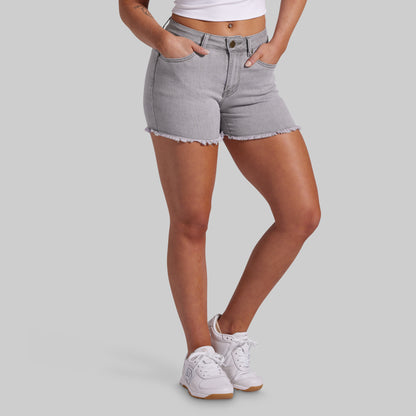 A model wearing light gray denim shorts with a frayed hem, paired with white sneakers and a white crop top, shown from the waist down on a solid gray background.