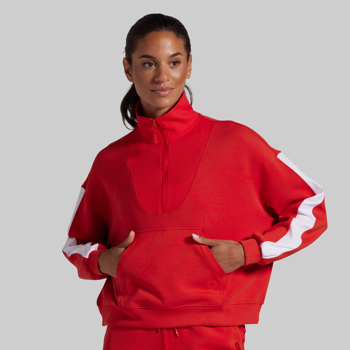 A woman modeling a bright red athletic half-zip sweatshirt with white contrast panels on the sleeves, paired with matching red pants, against a solid gray studio background.